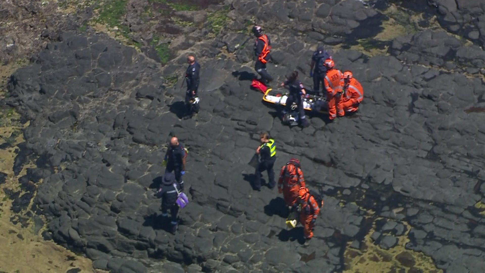 Una vista aérea de personas con monos azul marino y naranja paradas sobre rocas negras alrededor de una camilla con una manta blanca.