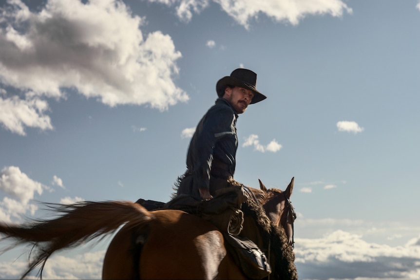 A scruffy 40-something man in a cowboy hat is riding a horse, but has his gaze turned behind him to look at the camera