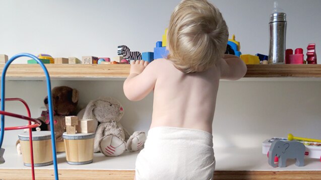 A baby wearing a cloth nappy stands at his bookcase of toys