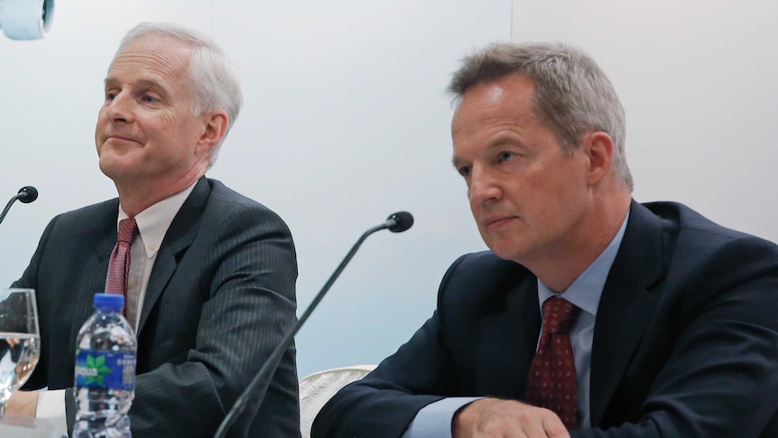 Rupert Hogg and John Slosar wear suits and sit in front of two microphones at a press conference