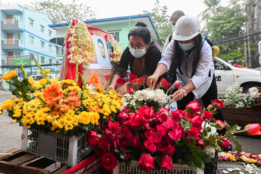 Two Asian women with face masks place flowers on colourful memorial with man's photo displayed.