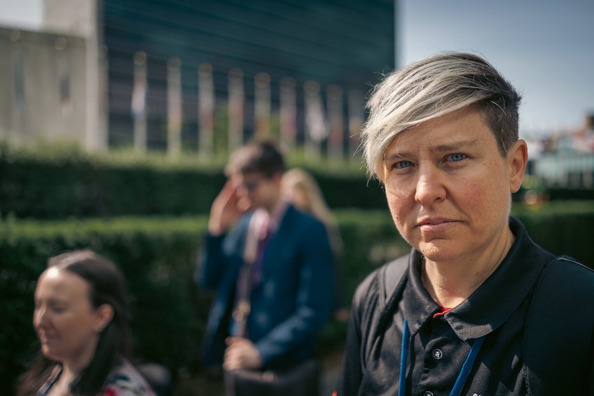 A woman looks into the camera with a serious expression. Behind her is the UN building. A group of people are next to her.