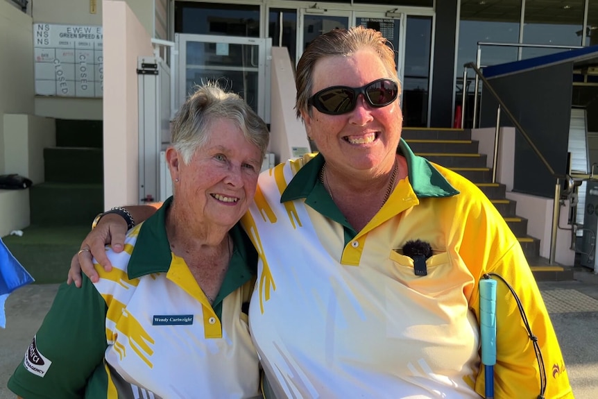 Two women stand with their arms around each other, smiling, wearing Australian sports shirts.