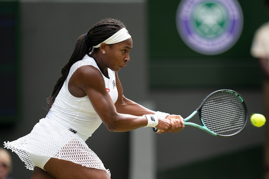A woman with dark braids wearing white tennis outfit hits the ball with her racquet.