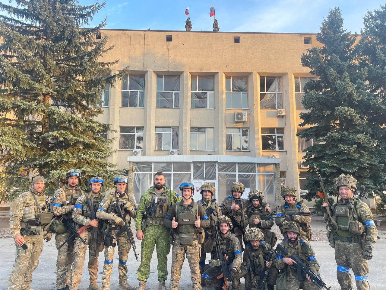 Ukrainian soldiers pose outside of building with Russian flags still flying above.