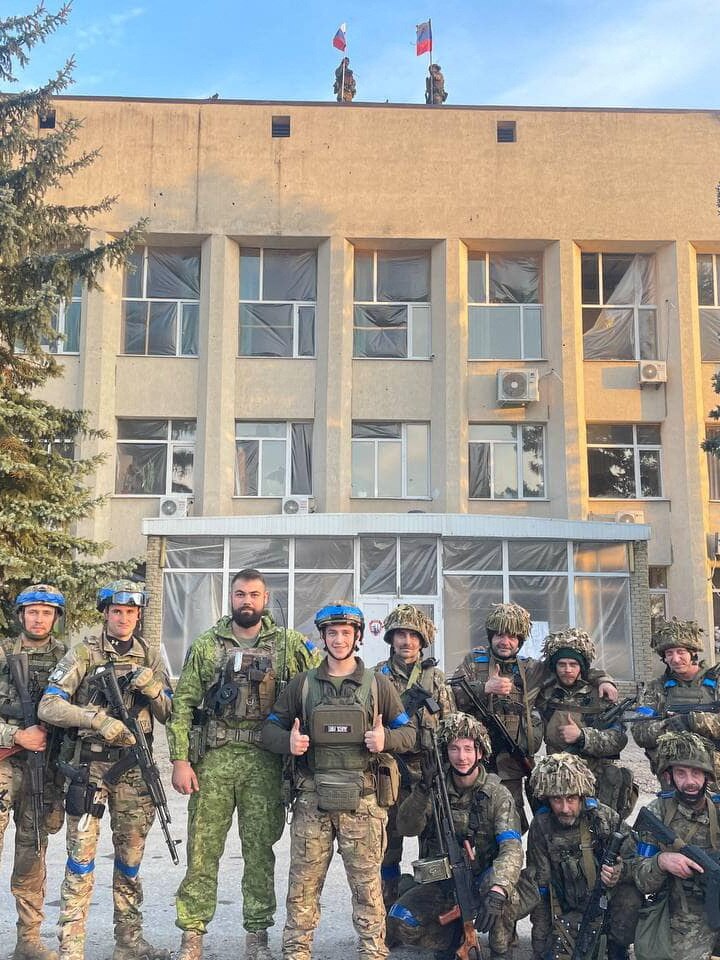 Ukrainian soldiers pose outside of building with Russian flags still flying above.