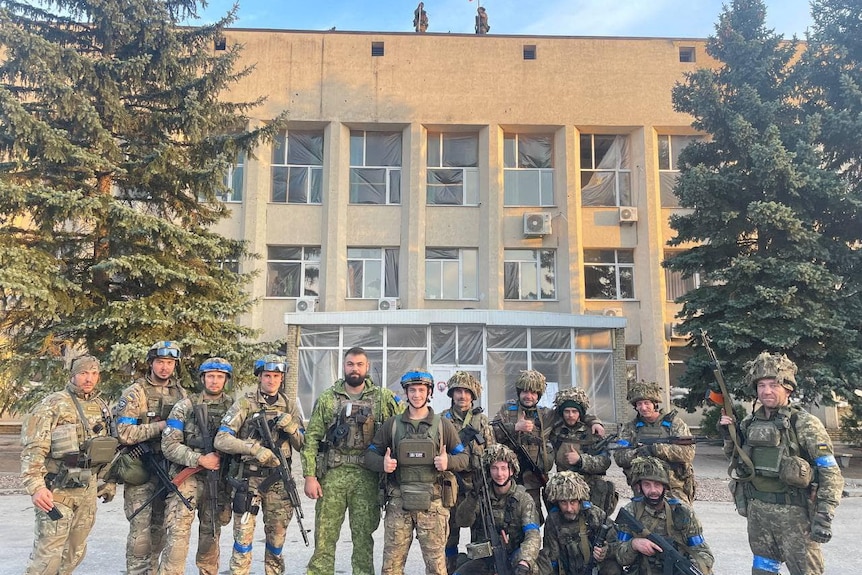 Ukrainian soldiers pose outside of building with Russian flags still flying above.