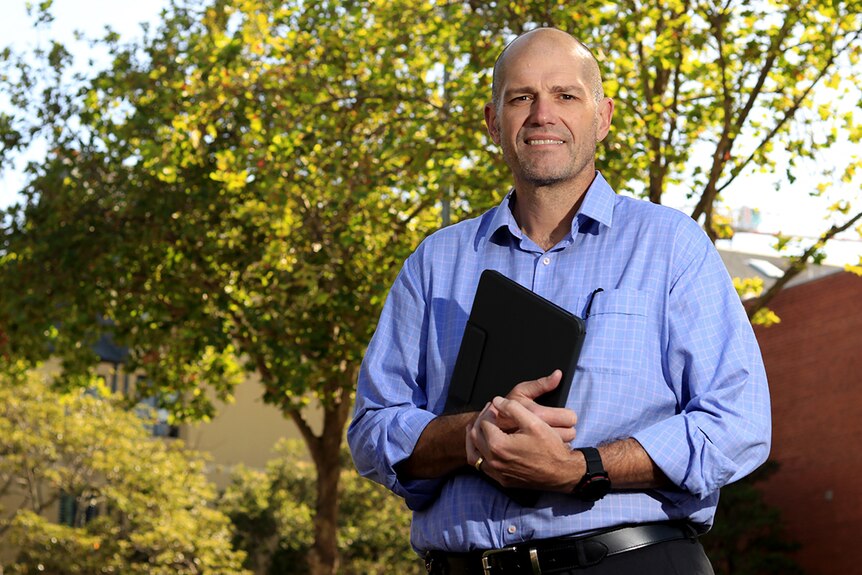 A man in a business shirt holds an electronic tablet outside under some trees.