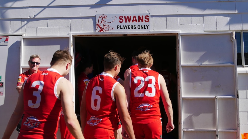 Three AFL players in red uniforms enter a dark room under a banner reading: Swans players only