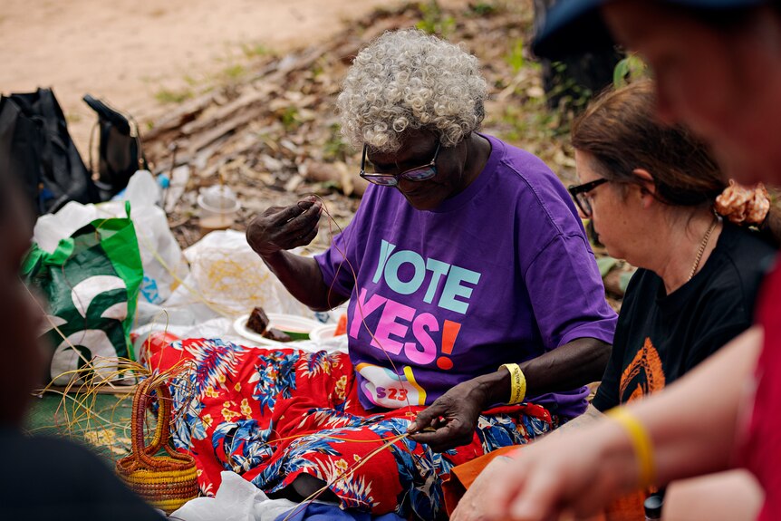 A woman in a purple t-shirt reading vote yes weaving a basket while surrounded by others