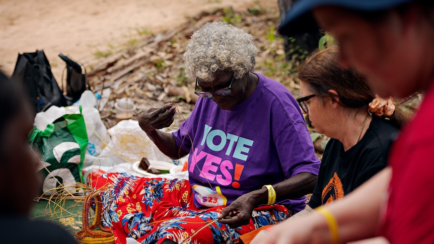 A woman in a purple t-shirt reading vote yes weaving a basket while surrounded by others