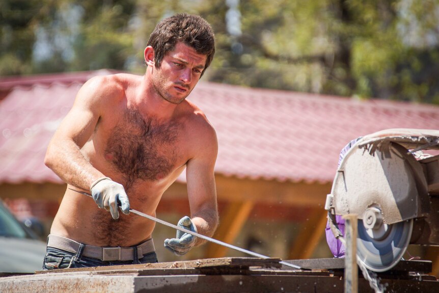 A topless worker in jeans and gloves cuts metal in full sun on a building site.