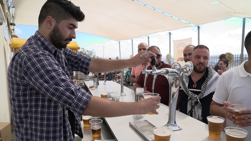 A man in a plaid shirt pours a beer as other ben stand waiting behind a bar