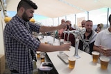 A man in a plaid shirt pours a beer as other ben stand waiting behind a bar