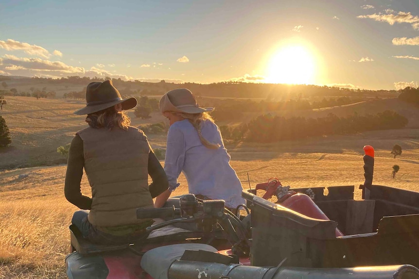 Two woman sit on the hood of an ATV looking out to the sunset. 