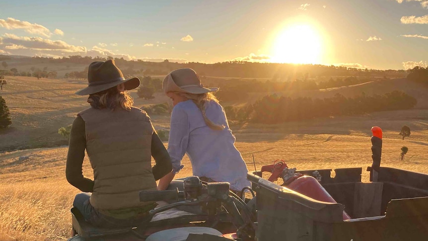 Two woman sit on the hood of an ATV looking out to the sunset. 