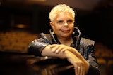 Deborah Cheetham Fraillon, with short, white hair stands behind a piano in Sydney Conservatorium of Music's Verbrugghen Hall.
