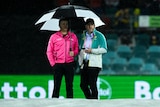 Two cricket match officials stand under an umbrella at Manuka Oval.