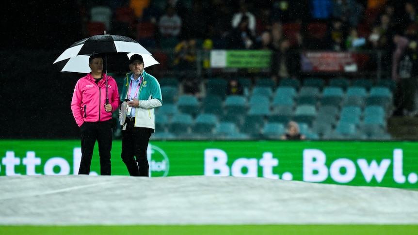 Two cricket match officials stand under an umbrella at Manuka Oval.