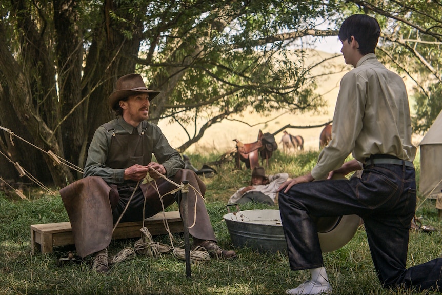 A teenage boy kneels in front of a 40-something man in a cowboy hat who is sitting braiding rope at a campsite