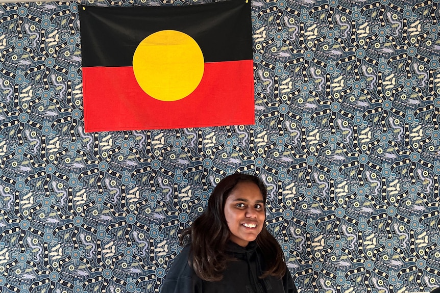 A teenager in a black jumper stands in front of a blue tapestry and an Aboriginal flag. 