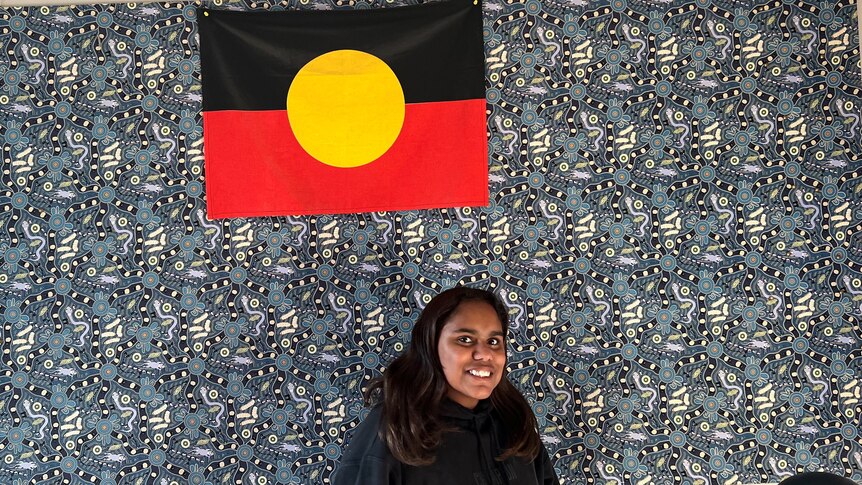 A teenager in a black jumper stands in front of a blue tapestry and an Aboriginal flag. 
