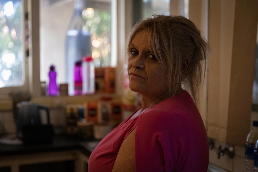 A woman with a weathered face, and a defeated expression, stands in a cluttered kitchen.