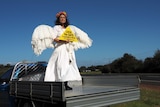 A woman dressed as an angel stands in the back of a flat-bed truck holding a sign saying 'lock the gate'.