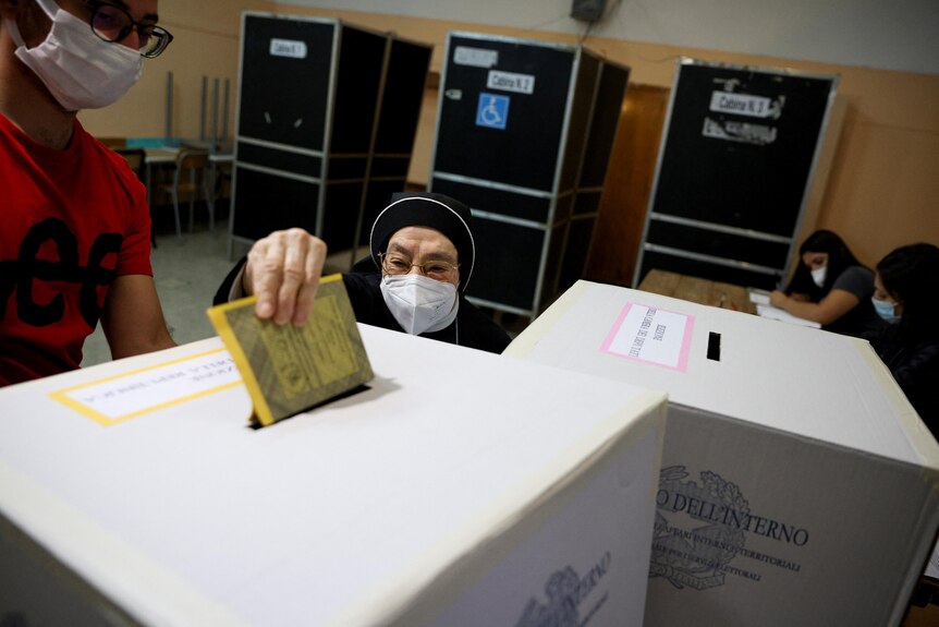 An elderly nun in a COVID mask drops a ballot into a box.