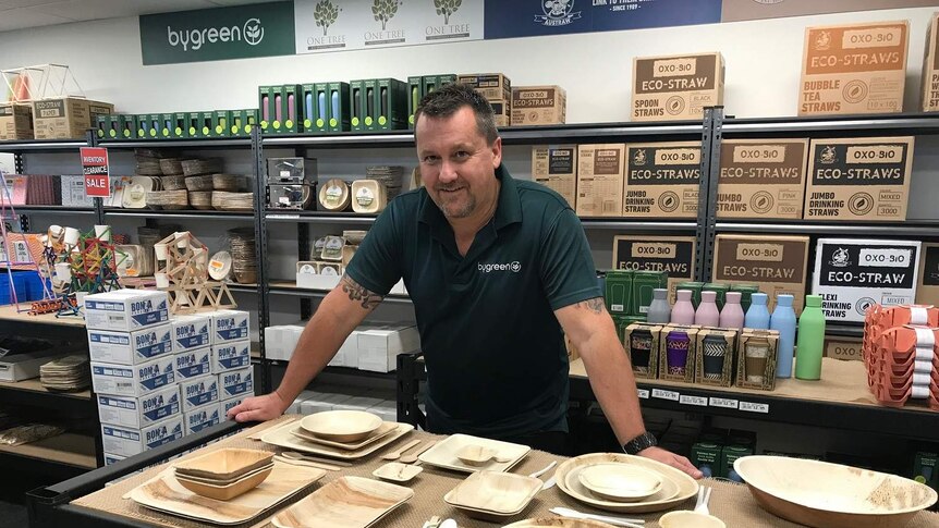 Mr Krause stands behind a counter which shows his eco plates and cutlery, behind him is eco straws.