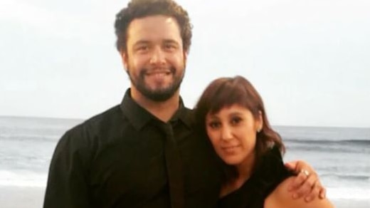 A smiling young man in a black shirt with his arm around a young woman. They are standing on a beach.