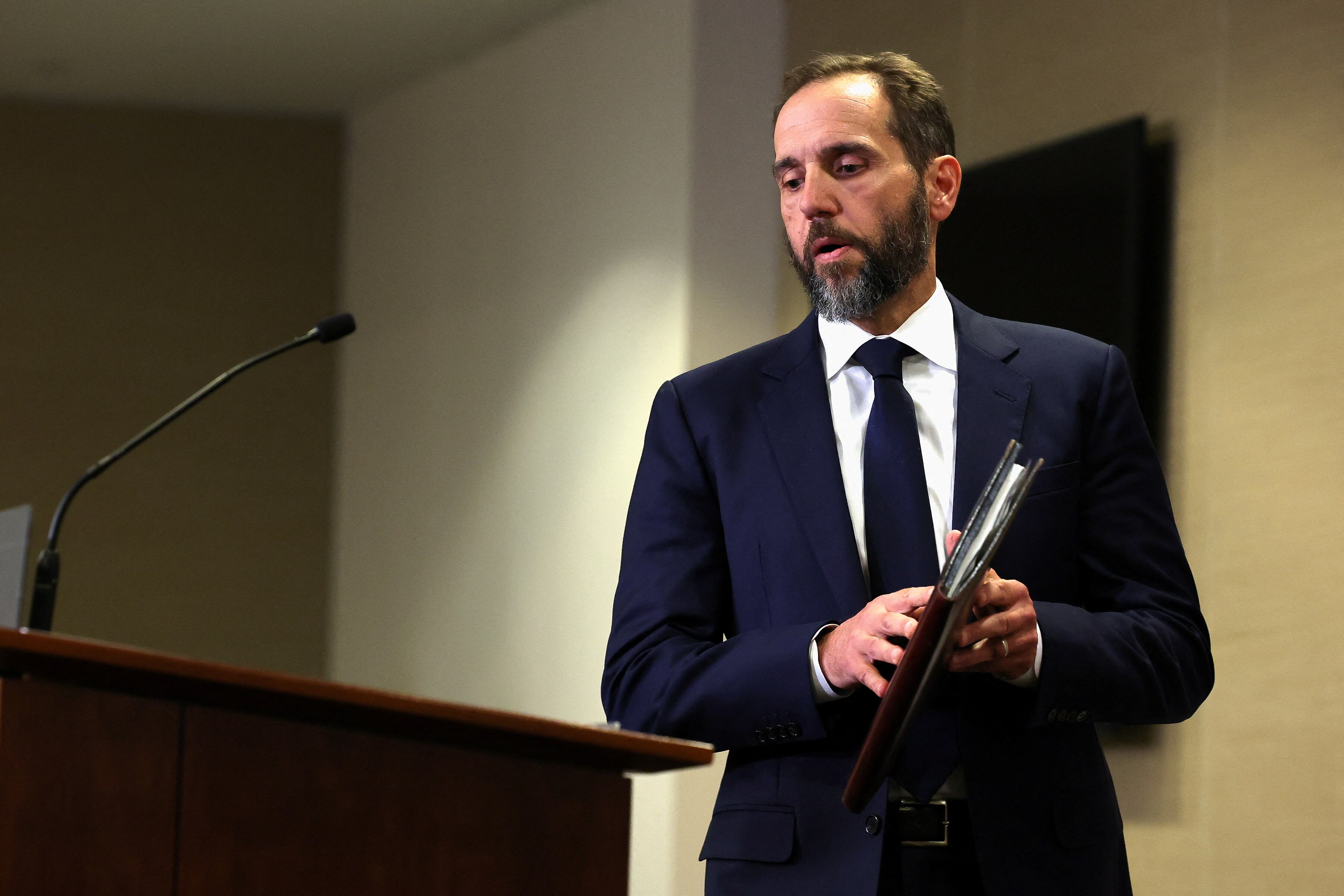 Jack Smith, wearing a dark navy suit and tie, holds a leatherbound folder as he approaches a microphone on a lectern