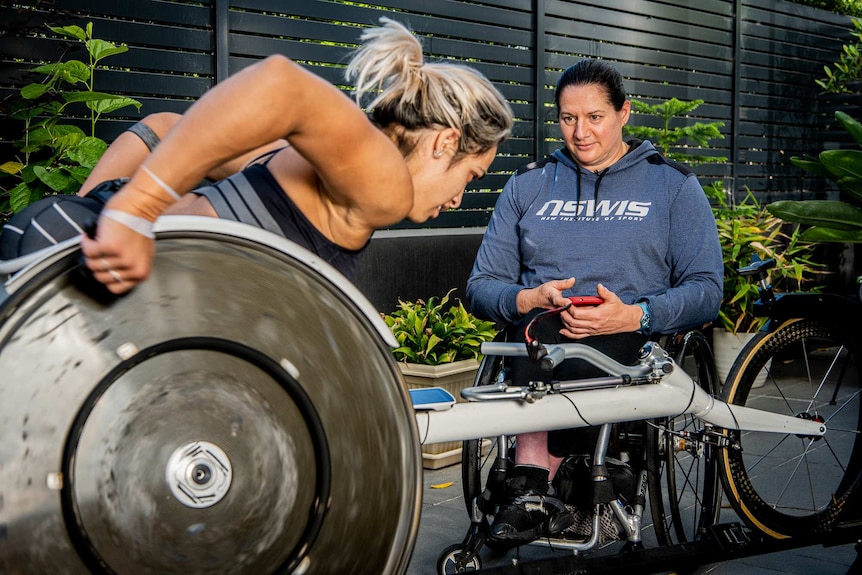 A woman in a wheelchair watches a younger woman in a racing wheelchair intently.