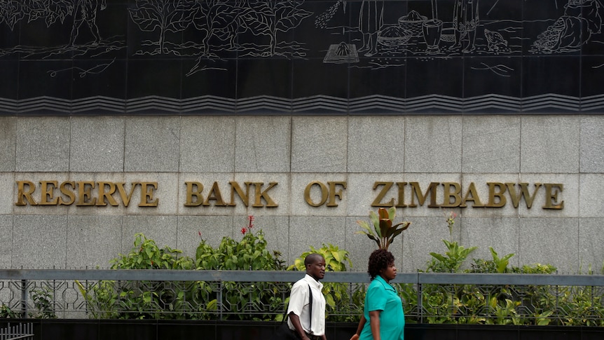 A man and a woman walk past a building made with large grey stones. In gold lettering reads 'reserve bank of zimbabwe'