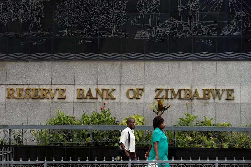 A man and a woman walk past a building made with large grey stones. In gold lettering reads 'reserve bank of zimbabwe'