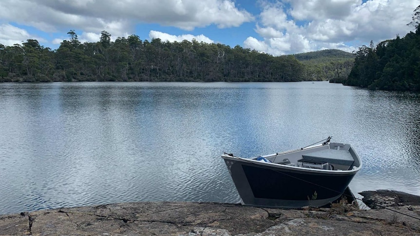 A boat sits on the rocky shore of a calm lake. There is a thick tree line against a cloudy horizon