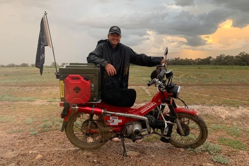 A man in a poncho stands smiling next to a red and black honda motorbike.
