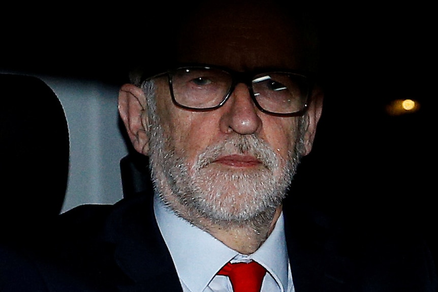 Jeremy Corbyn, partially in shadow, frowns while wearing a rosette