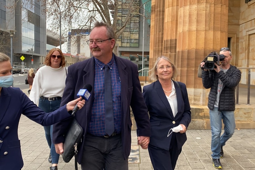 A man and a woman, both wearing blue suits, smile as they hold hands and walk outside a court building