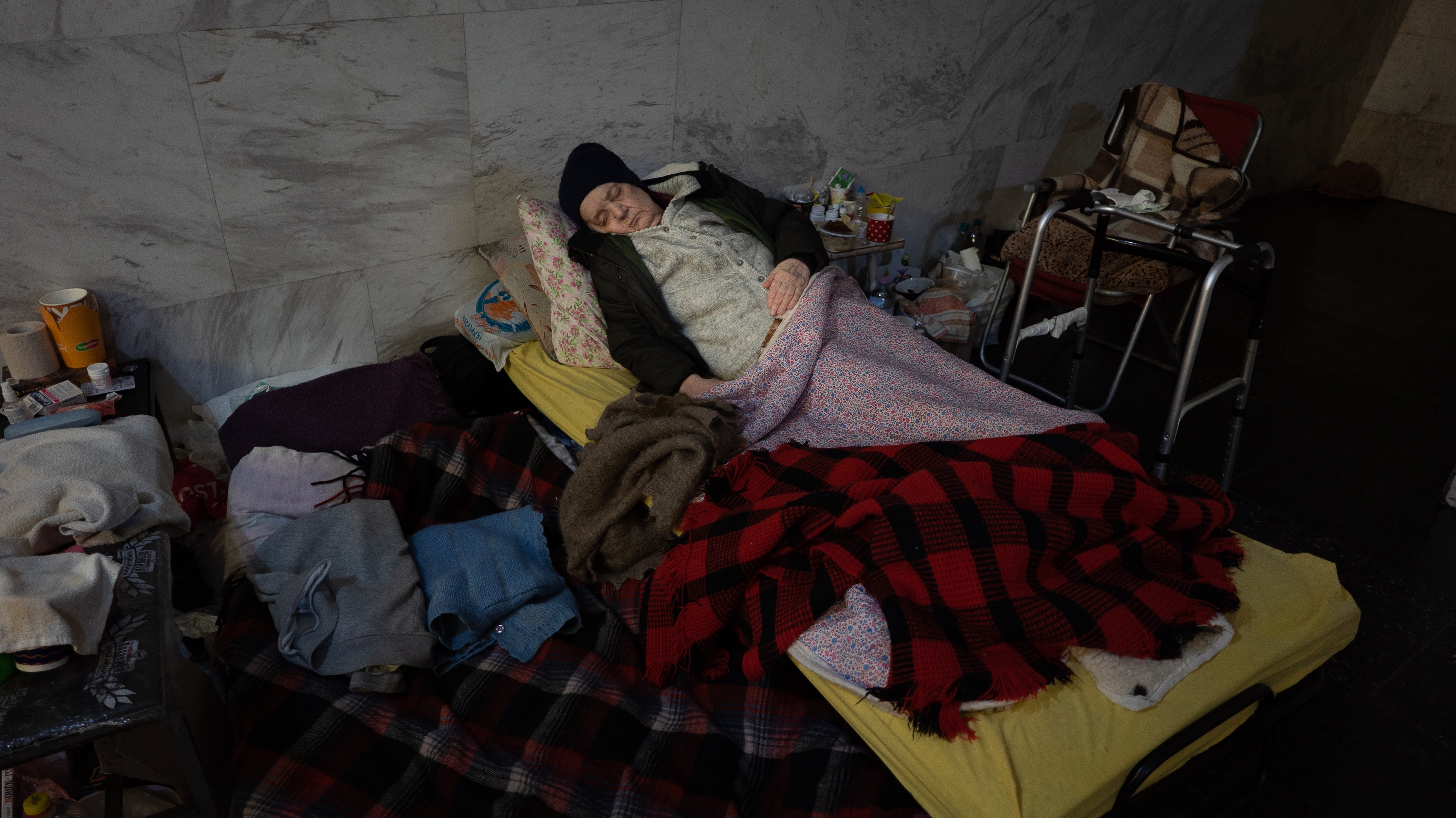 An older woman sleeps on a pile of blankets with her walker next to her. 