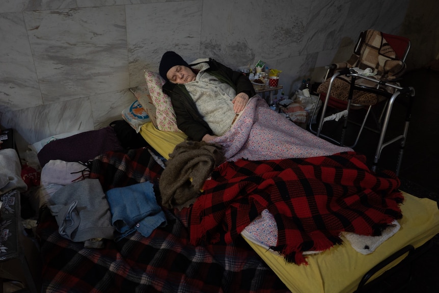 An older woman sleeps on a pile of blankets with her walker next to her. 