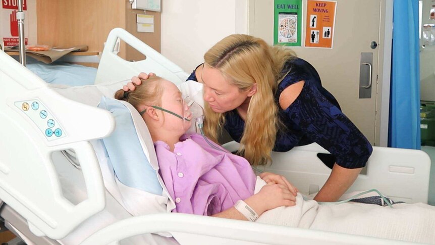 Ms Stuart lays in a hospital bed with an oxygen mask, as a nurse looks on.