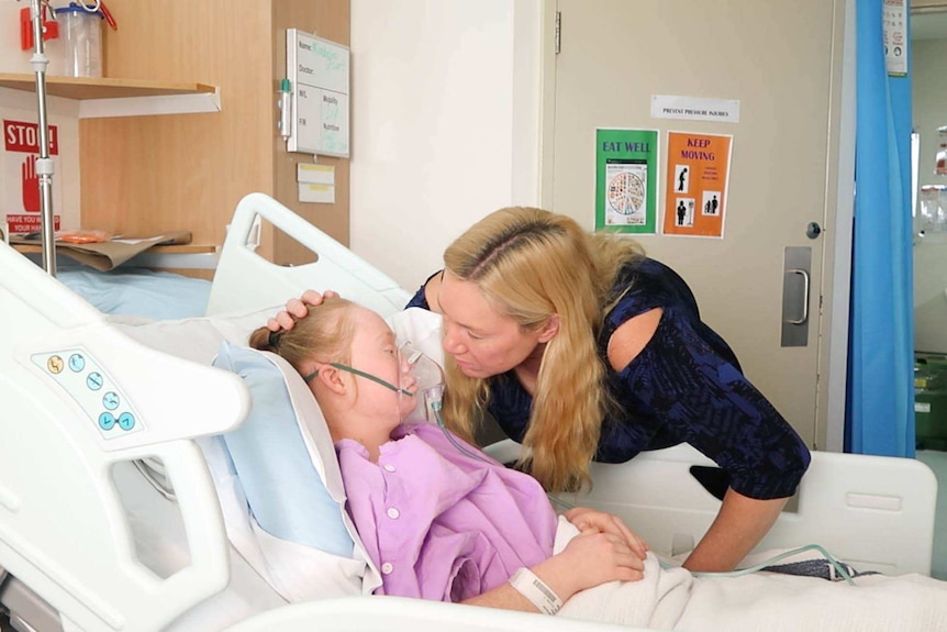 Ms Stuart lays in a hospital bed with an oxygen mask, as a nurse looks on.