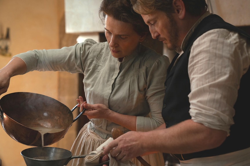 A film still of Juliette Binoche and Benoît Magimel, both in late 19th century dress, pouring liquid between two pans.
