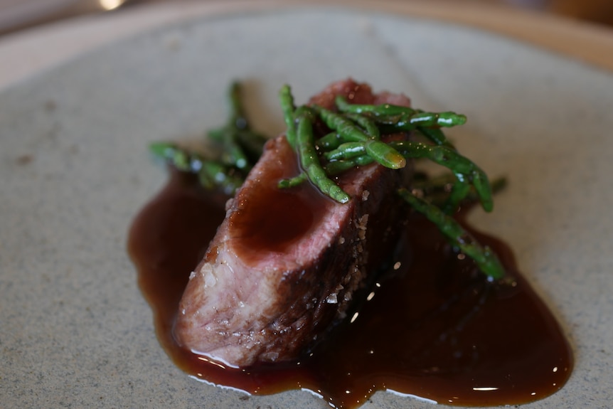 a close shot of medium rare vension with a gravy sauce poured over and around it, and samphire greens on top.