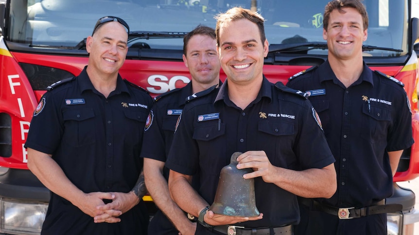 Four firefighters stand before a truck, one stands in the front holding an aged bell.