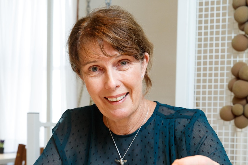 A smiling middle-aged woman  with brown-reddish hair, blue dotted top, chain around neck, sits in a room with white mesh behind.