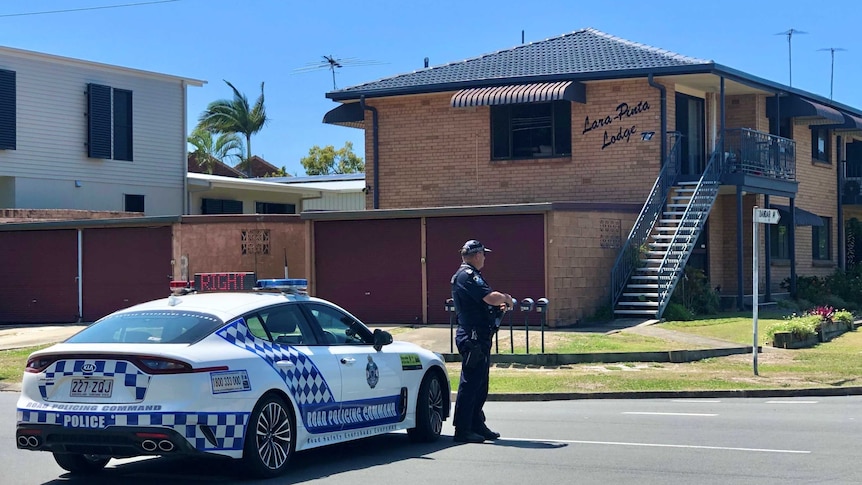 A police officer stands on the road in front of a white police vehicle, looking over to brick units.