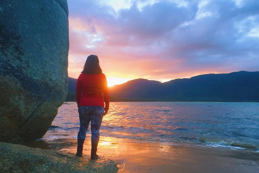 A woman stands on a beach, facing away from the camera, looking at a colourful sunset.