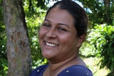 A Fijian woman standing side on smiles with trees in the background